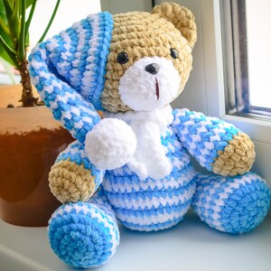 May include: A crocheted teddy bear wearing a blue and white striped sleepwear set with a white pom-pom on the hat. The bear is sitting on a windowsill.