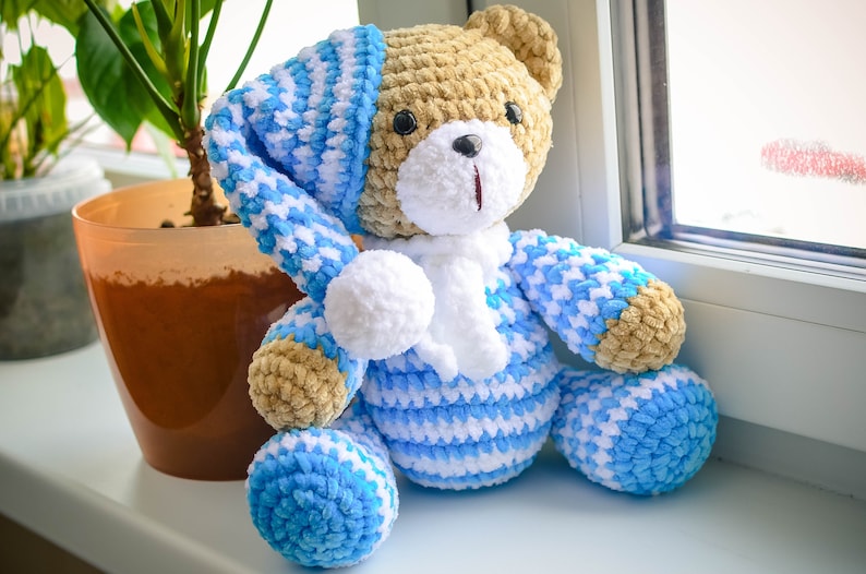 May include: A crocheted teddy bear wearing a blue and white striped sleepwear set with a white pom-pom on the hat. The bear is sitting on a windowsill.