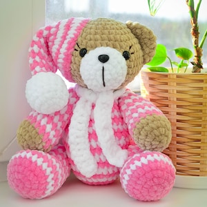 May include: A crocheted teddy bear wearing a pink and white striped sleep cap and a white scarf. The bear is sitting on a windowsill with a potted plant in the background.