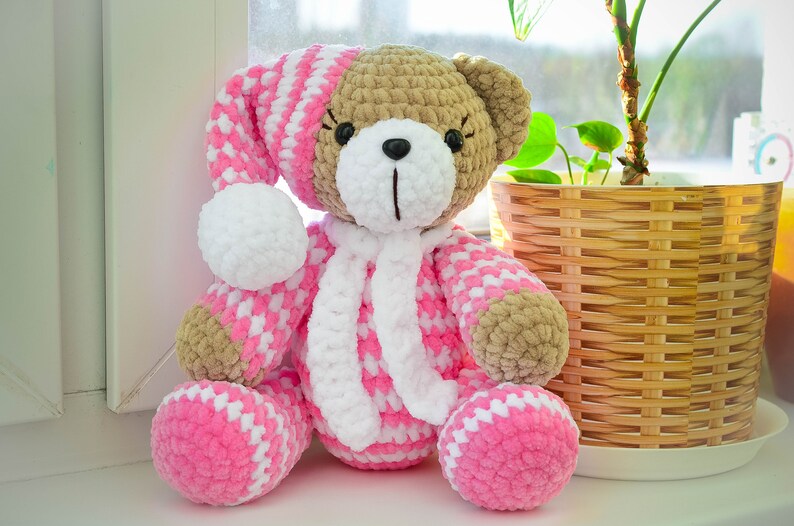 May include: A crocheted teddy bear wearing a pink and white striped sleep cap and a white scarf. The bear is sitting on a windowsill with a potted plant in the background.