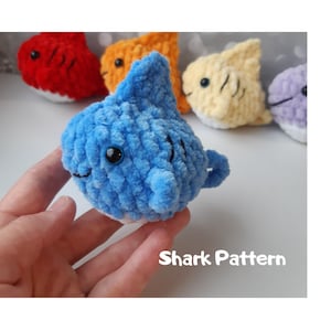 May include: A blue crocheted shark toy with black eyes and a white belly. The toy is being held in a hand. The text "Shark Pattern" is visible below the toy.