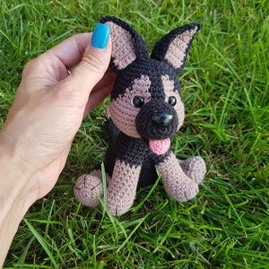 May include: A crocheted stuffed dog toy with black and brown fur, brown eyes, a pink tongue, and black nose. The toy is sitting on a bed of green grass.