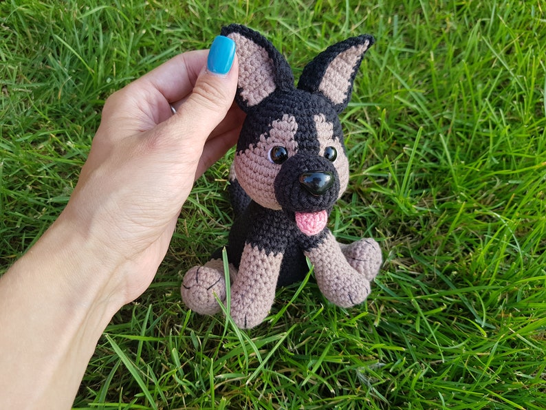 May include: A crocheted stuffed dog toy with black and brown fur, brown eyes, a pink tongue, and black nose. The toy is sitting on a bed of green grass.