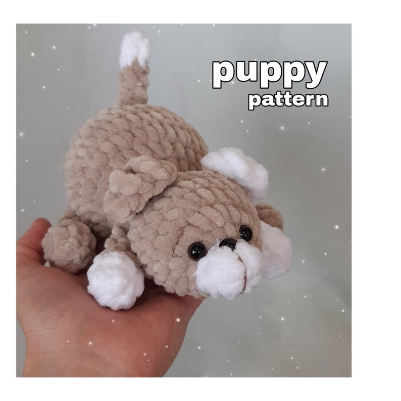 May include: A crocheted beige puppy toy with white accents on the ears, paws, and tail. The toy is held in a person's hand. The text "puppy pattern" is written in black above the toy.
