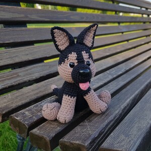 May include: A crocheted plush toy dog sitting on a wooden bench. The dog is black and brown with a pink tongue sticking out.