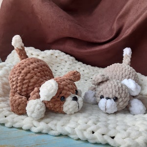 May include: Two crocheted plush dog toys, one brown and one gray, lying on a white fuzzy blanket. The dogs have white ears and noses.