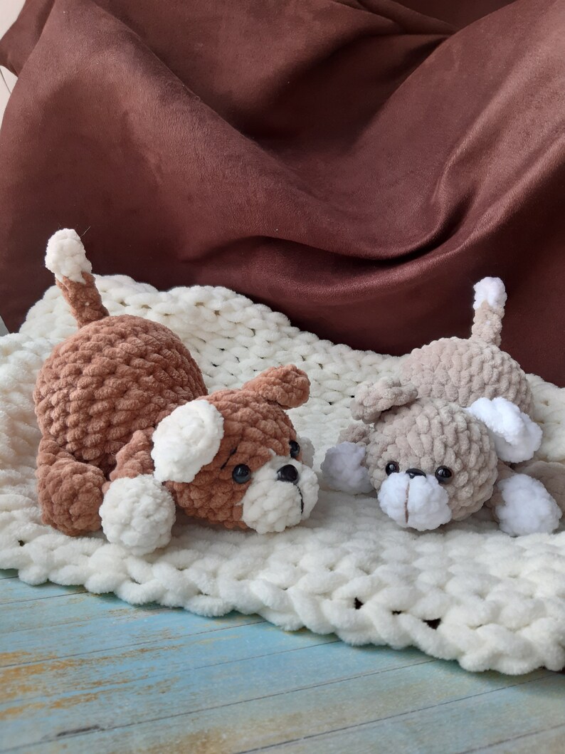 May include: Two crocheted plush dog toys, one brown and one gray, lying on a white fuzzy blanket. The dogs have white ears and noses.