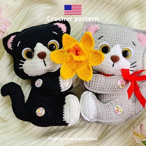 May include: Two handmade crochet cat plushies, one black and one gray, with white accents, button details, and large, expressive eyes. A yellow flower is placed between them. The text "Crochet pattern" and "English (American terms)" are visible.