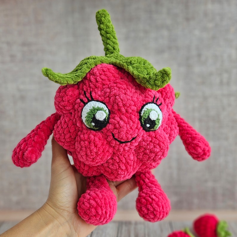 May include: A crocheted plush toy shaped like a raspberry with green leaves and a smiling face. The toy has large, white eyes with green pupils and a pink, fuzzy body.