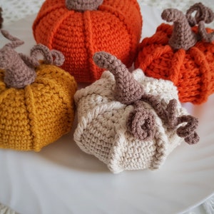 May include: Three crocheted pumpkins in orange, yellow, and white. The pumpkins are sitting on a white surface.