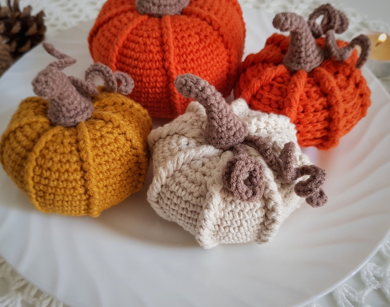 May include: Three crocheted pumpkins in orange, yellow, and white. The pumpkins are sitting on a white surface.