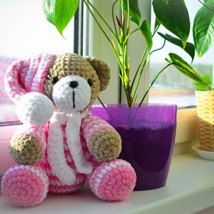 May include: A crocheted teddy bear wearing a pink and white striped hooded outfit sits on a windowsill with two potted plants.