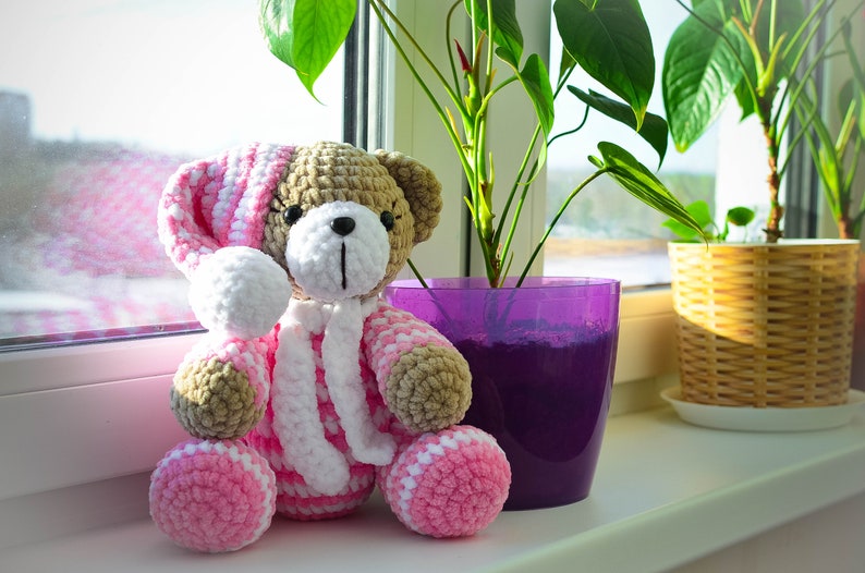 May include: A crocheted teddy bear wearing a pink and white striped hooded outfit sits on a windowsill with two potted plants.