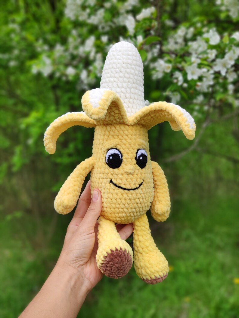 May include: A crocheted banana plush toy with a smiling face and brown feet. The banana is yellow with a white crocheted top.