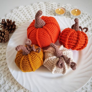 May include: Four crocheted pumpkins in orange, white, and yellow on a white doily. The pumpkins are arranged in a circle on a white plate.