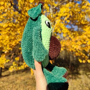 K&ouml;nnte beinhalten: Ein gr&uuml;nes und braunes geh&auml;keltes Avocado-Pl&uuml;schtier mit einem Blatt oben. Das Spielzeug wird in einer Hand vor einem Hintergrund aus gelben Herbstbl&auml;ttern gehalten.