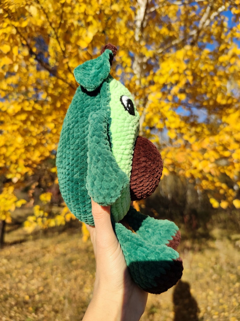 K&ouml;nnte beinhalten: Ein gr&uuml;nes und braunes geh&auml;keltes Avocado-Pl&uuml;schtier mit einem Blatt oben. Das Spielzeug wird in einer Hand vor einem Hintergrund aus gelben Herbstbl&auml;ttern gehalten.