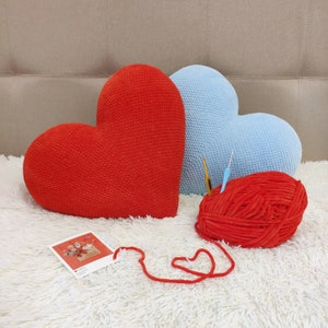 May include: Two heart-shaped pillows, one red and one light blue, are on a white fluffy surface. A ball of red yarn is in front of the pillows.