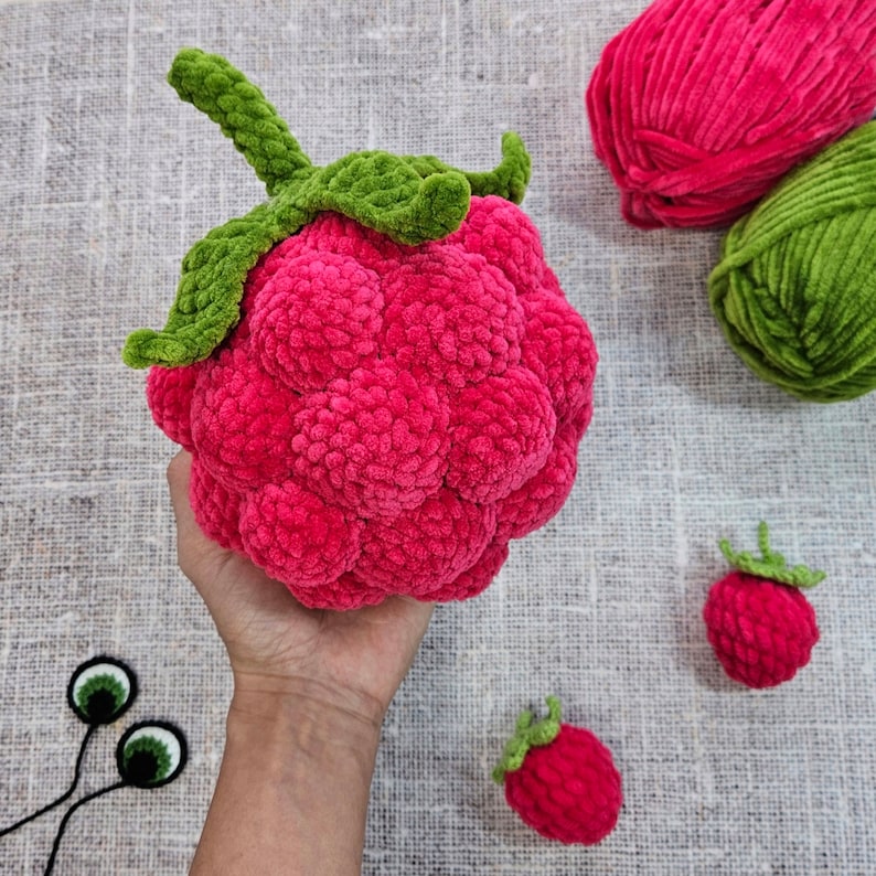 May include: A hand holds a large crocheted raspberry with green leaves. Two smaller crocheted raspberries are on a textured surface.