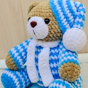 May include: A crocheted teddy bear wearing a blue and white patterned hat and matching pajamas. The bear has brown fur and a white face with black eyes and a brown nose.