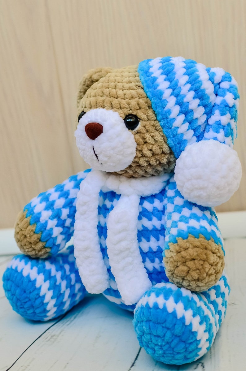May include: A crocheted teddy bear wearing a blue and white patterned hat and matching pajamas. The bear has brown fur and a white face with black eyes and a brown nose.