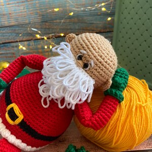 May include: A crocheted Santa Claus doll with a white beard, red and white hat, and a black belt. The doll is lying on its side with a yellow ball of yarn.