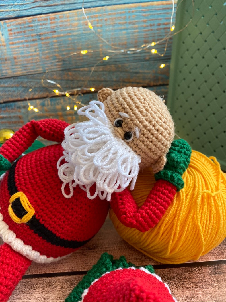 May include: A crocheted Santa Claus doll with a white beard, red and white hat, and a black belt. The doll is lying on its side with a yellow ball of yarn.