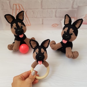 K&ouml;nnte beinhalten: Drei geh&auml;kelte Sch&auml;ferhund-Pl&uuml;schtiere, eines davon mit einem Holz-Bei&szlig;ring. Die Pl&uuml;schtiere sind braun und schwarz mit rosa Nasen und Zungen. Ein Pl&uuml;schtier h&auml;lt einen roten Ball.