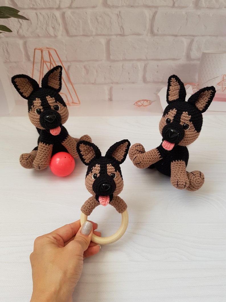 K&ouml;nnte beinhalten: Drei geh&auml;kelte Sch&auml;ferhund-Pl&uuml;schtiere, eines davon mit einem Holz-Bei&szlig;ring. Die Pl&uuml;schtiere sind braun und schwarz mit rosa Nasen und Zungen. Ein Pl&uuml;schtier h&auml;lt einen roten Ball.