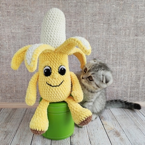 May include: A crocheted yellow banana toy with a smiling face sits on a green stool. A gray tabby kitten looks at the banana toy.