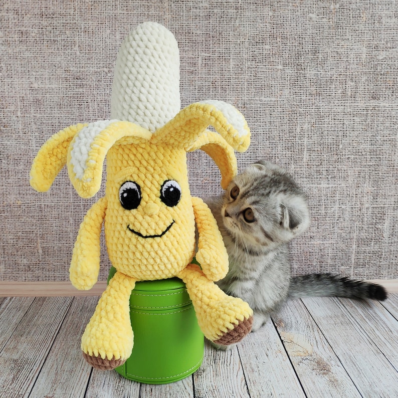 May include: A crocheted yellow banana toy with a smiling face sits on a green stool. A gray tabby kitten looks at the banana toy.