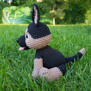 May include: A crocheted stuffed dog toy in black and brown, sitting on green grass. The dog has a pink tongue sticking out.