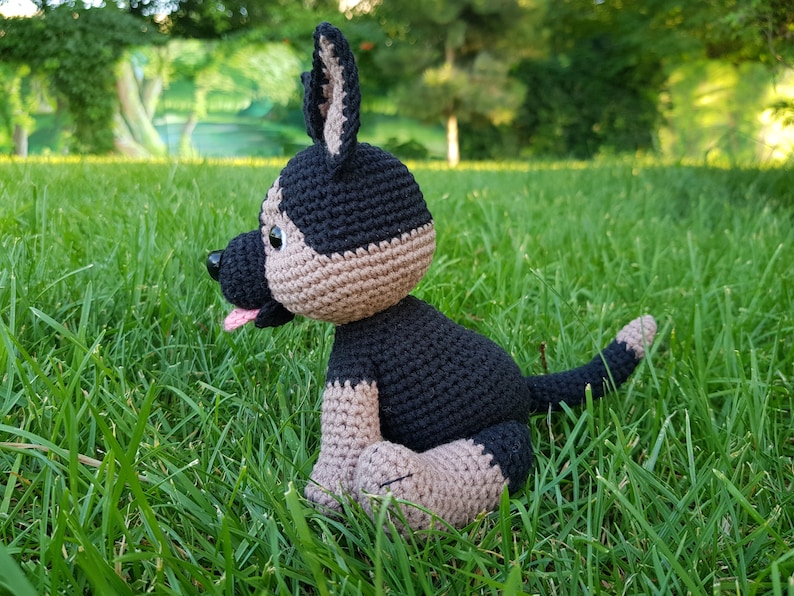 May include: A crocheted stuffed dog toy in black and brown, sitting on green grass. The dog has a pink tongue sticking out.