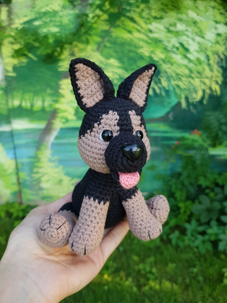 K&ouml;nnte beinhalten: Ein geh&auml;keltes Stofftier in Form eines Hundes mit schwarzem und braunem Fell, schwarzen Augen und einer rosa Zunge. Der Hund sitzt mit gekreuzten Beinen.