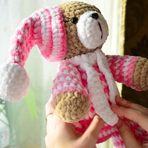 May include: A crocheted teddy bear wearing a pink and white crocheted hat and matching pajamas. The bear is being held by a hand.