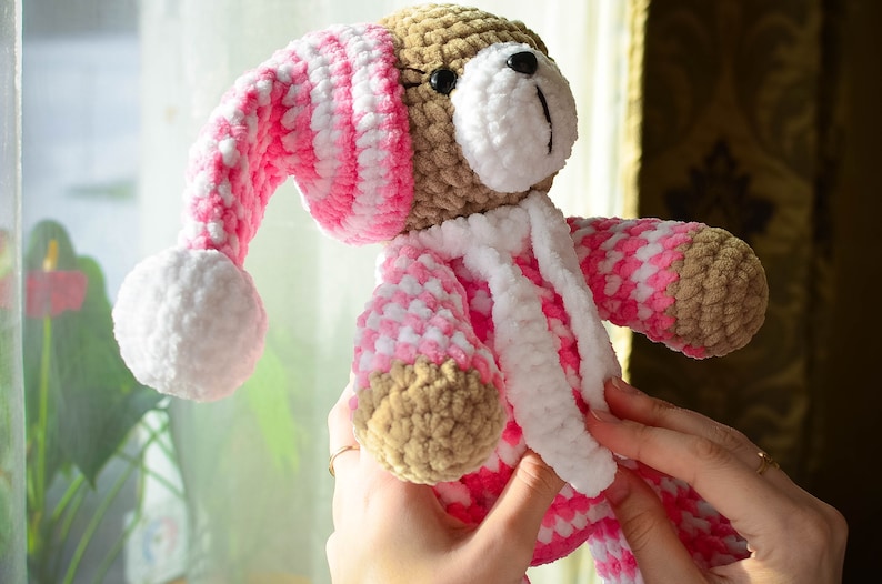 May include: A crocheted teddy bear wearing a pink and white crocheted hat and matching pajamas. The bear is being held by a hand.