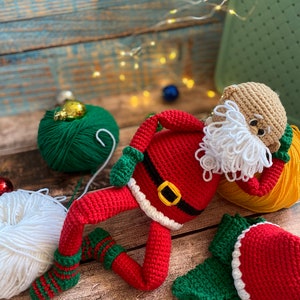May include: A crocheted Santa Claus doll with a white beard, red suit, and green hat. The doll is lying on a wooden surface with yarn balls in the background.