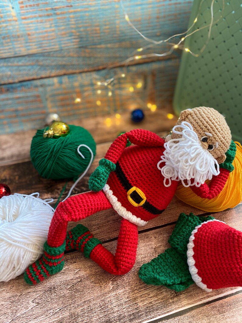 May include: A crocheted Santa Claus doll with a white beard, red suit, and green hat. The doll is lying on a wooden surface with yarn balls in the background.