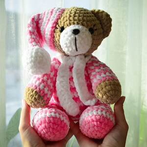 May include: A crocheted teddy bear wearing a pink and white striped sleep cap and matching pajamas. The bear is brown and has black eyes and a black nose.