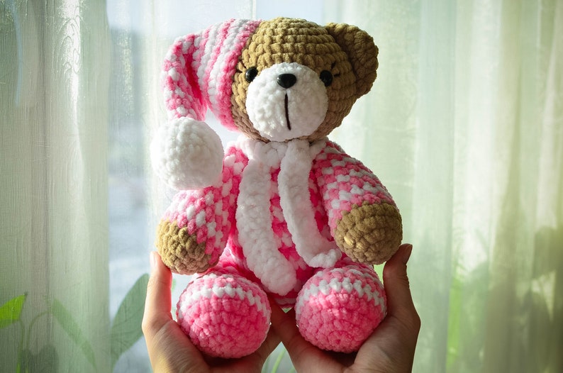 May include: A crocheted teddy bear wearing a pink and white striped sleep cap and matching pajamas. The bear is brown and has black eyes and a black nose.