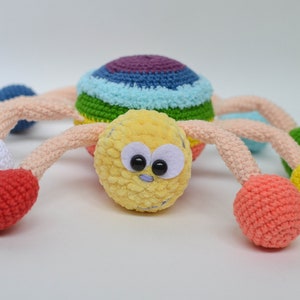 Pu&ograve; includere: Un giocattolo ragno all'uncinetto colorato con una testa gialla, occhi neri e gambe arcobaleno. Le gambe sono sormontate da palline all'uncinetto di vari colori, tra cui viola, bianco, rosso, verde e giallo.