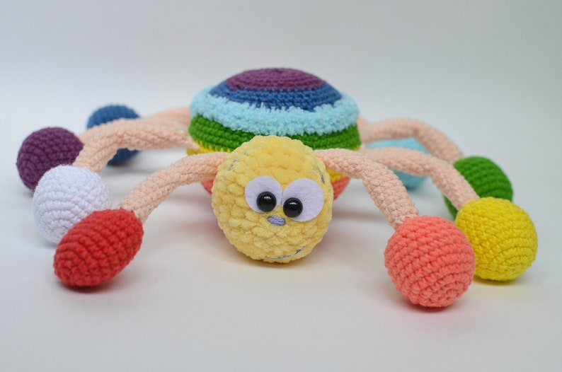 Pu&ograve; includere: Un giocattolo ragno all'uncinetto colorato con una testa gialla, occhi neri e gambe arcobaleno. Le gambe sono sormontate da palline all'uncinetto di vari colori, tra cui viola, bianco, rosso, verde e giallo.