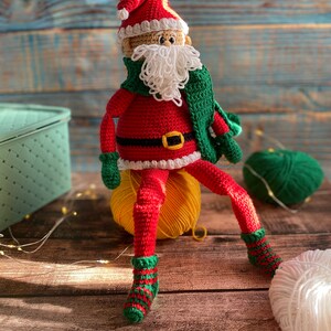 May include: A crocheted Santa Claus doll wearing a red suit, white beard, green scarf, and green and red striped socks. The doll is sitting on a yellow yarn ball.