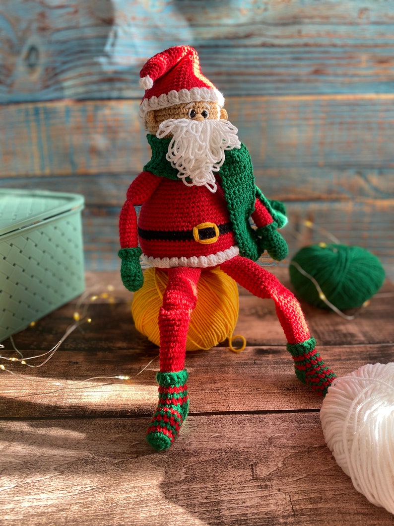 May include: A crocheted Santa Claus doll wearing a red suit, white beard, green scarf, and green and red striped socks. The doll is sitting on a yellow yarn ball.