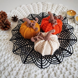 May include: Three crocheted pumpkins in orange, beige, and yellow, sitting on a black crocheted doily on a white crocheted surface.