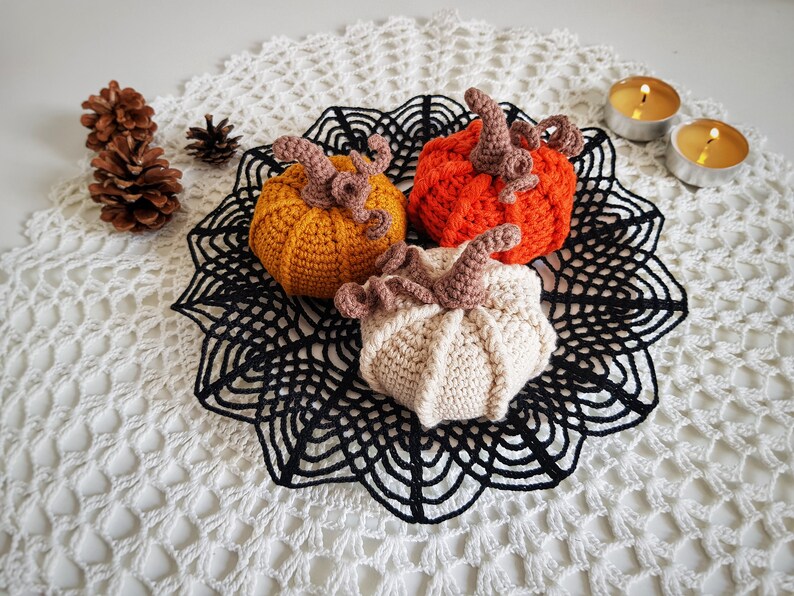 May include: Three crocheted pumpkins in orange, beige, and yellow, sitting on a black crocheted doily on a white crocheted surface.