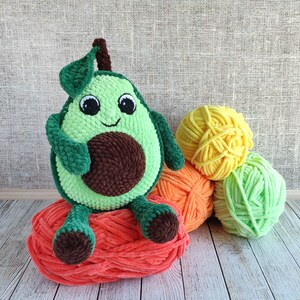 K&ouml;nnte beinhalten: Ein gr&uuml;nes, geh&auml;keltes Avocado-Pl&uuml;schtier mit einem l&auml;chelnden Gesicht und braunem Garn f&uuml;r den Kern. Die Avocado sitzt auf einem Stapel orangefarbenem, gelbem und gr&uuml;nem Garn.