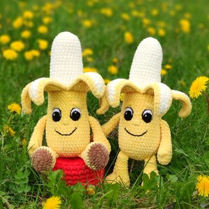 May include: Two crocheted banana plush toys with smiling faces, sitting in a field of green grass and yellow flowers. One banana is sitting on a red crocheted cushion.