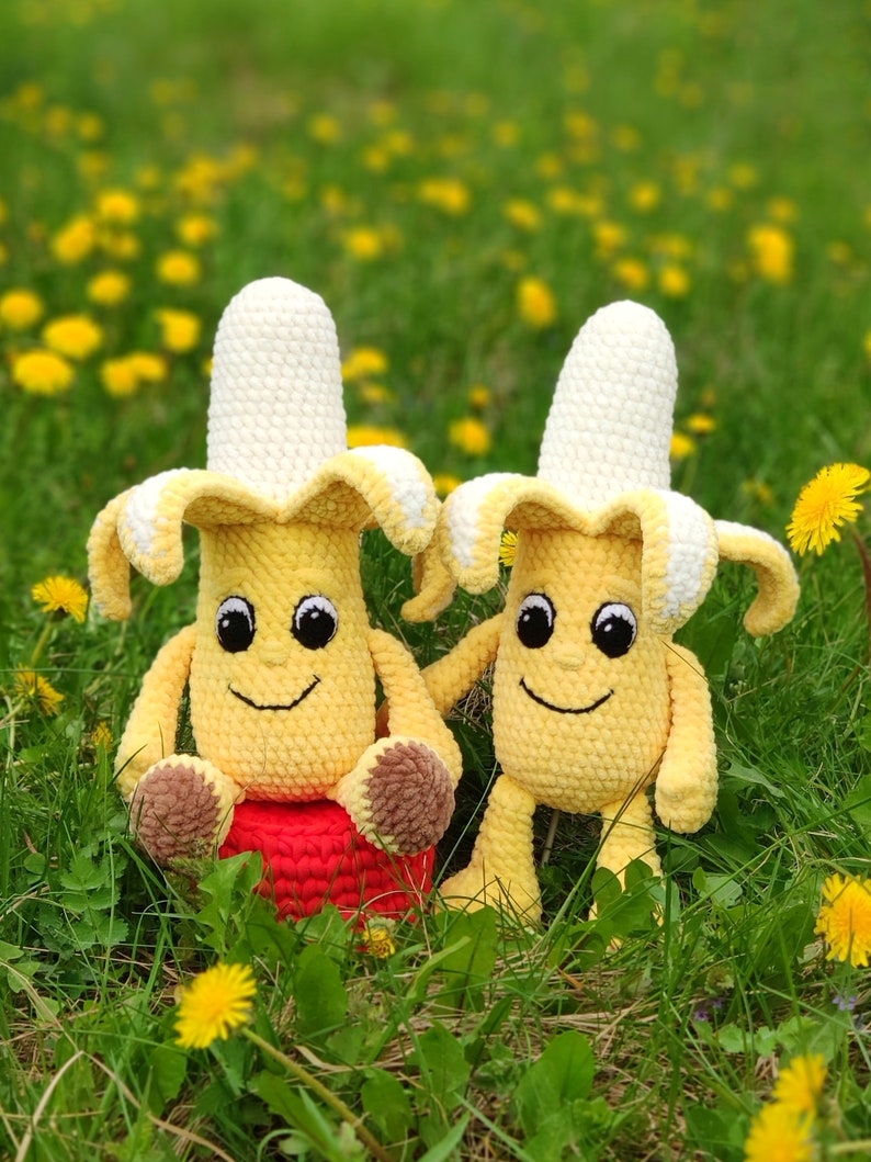 May include: Two crocheted banana plush toys with smiling faces, sitting in a field of green grass and yellow flowers. One banana is sitting on a red crocheted cushion.