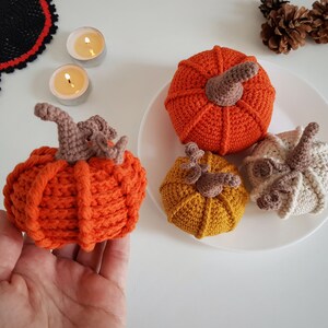 May include: Four crocheted pumpkins in orange, yellow, and cream with brown stems. The pumpkins are on a white plate.